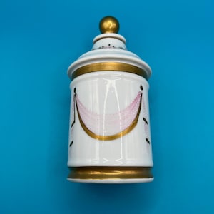 May include: White ceramic jar with a lid, featuring gold trim and a gold knob on top. The jar is decorated with pink and gold accents. The jar is on a blue background.