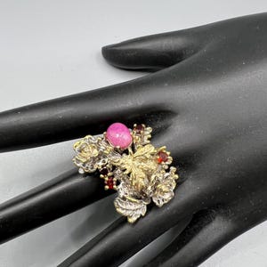 May include: A vintage ring with a floral design. The ring features a large, oval, pink gemstone, surrounded by smaller red gemstones and gold and silver-toned metal floral accents. The ring is set on a black surface.