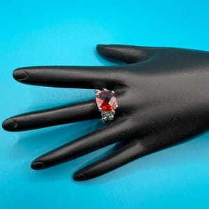 May include: A silver ring with a large, faceted, red gemstone. The gemstone is oval-shaped and set in a silver band with decorative accents. The ring is displayed on a black hand against a bright blue background.