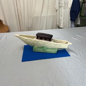 May include: Cream-colored ceramic boat-shaped planter with a dark brown rectangular accent. The planter has a light green base with scroll details and is set on a blue surface. The boat has a ribbed design.