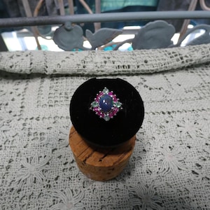 May include: A silver ring with a large oval blue gemstone at the center, surrounded by smaller pink and green gemstones. The ring is displayed on a black velvet surface atop a cork.