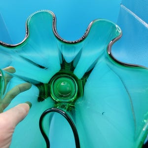 May include: A teal-colored glass bowl with a unique, organic shape. The bowl has a dark green center and a silver-colored trim around the edges. The bowl's design resembles a flower or a star, with curved petals or points.