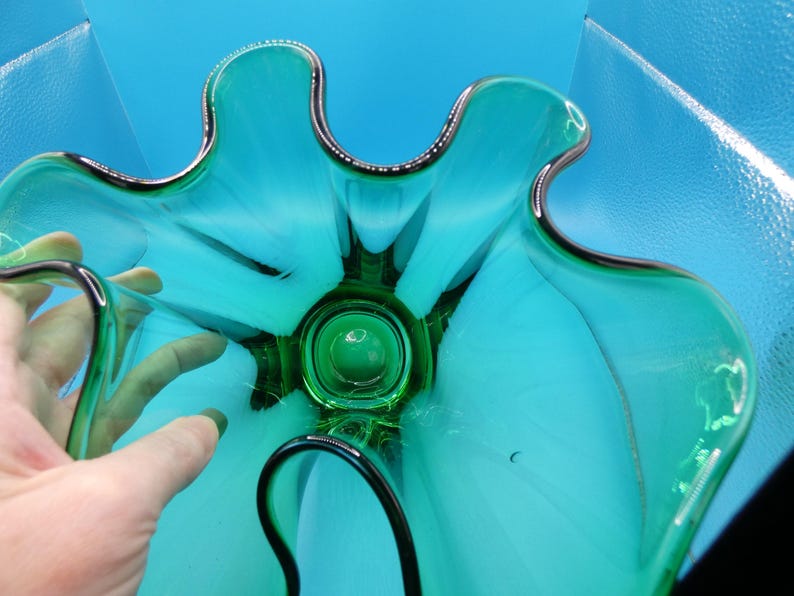 May include: A teal-colored glass bowl with a unique, organic shape. The bowl has a dark green center and a silver-colored trim around the edges. The bowl's design resembles a flower or a star, with curved petals or points.