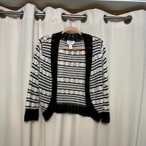 May include: A black and white striped cardigan sweater with a floral pattern. The sweater has a black collar, cuffs, and trim. The garment is hanging on a pink hanger against a white curtain backdrop.