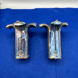 May include: Two ceramic vases with a unique, stylized design. The vases are primarily blue and brown with gold accents. Each vase has a flared top and a curved handle-like extension. The base of each vase features a floral design.