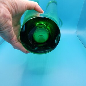 May include: A green glass bottle with two spouts, held by a hand. The bottle has a dark green base and a clear body. The background is a light blue.