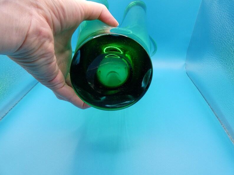 May include: A green glass bottle with two spouts, held by a hand. The bottle has a dark green base and a clear body. The background is a light blue.