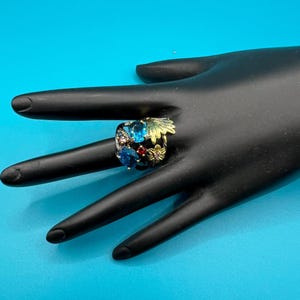 May include: A colorful ring with a floral design. The ring features blue, red, and pink gemstones, with gold and green accents. The ring is set on a black surface against a blue background.