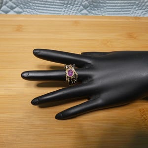 May include: A gold-toned ring with a central oval-shaped, pink gemstone. The ring has a detailed, textured design with small, dark accents. The ring is displayed on a black hand-shaped display.