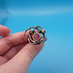 May include: A silver-toned ring with a central oval, pink gemstone, surrounded by a gold setting. The ring features small, round, turquoise-colored stones and an intricate, openwork design. The ring is held against a blue background.