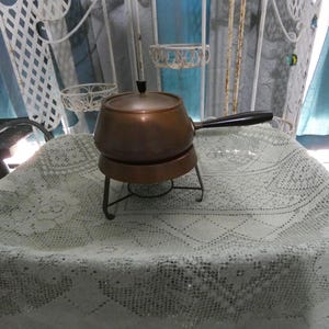 May include: A copper fondue pot with a black handle and a metal stand. The pot is on a white crocheted tablecloth.