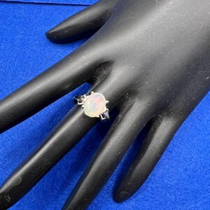 May include: A silver ring with an oval, iridescent opal gemstone. The opal has a mix of white, pink, and orange hues. The ring is set on a black surface with a blue background.