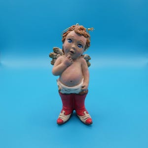 May include: A cherubic figurine of an angel with curly brown hair, blue eyes, and small gold wings. The angel is wearing red boots with white accents and a white diaper. The figurine is a decorative item.