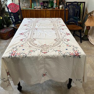 May include: Cream-colored rectangular tablecloth with intricate floral embroidery in pink, blue, and brown. The embroidery forms a central design and borders the edges. The tablecloth is draped over a black table.