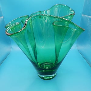 May include: A vibrant green glass vase with a ruffled rim and a flared, sculptural design. The vase has a rounded base and a wide opening, showcasing a modern aesthetic. The rim is accented with a thin gold trim.
