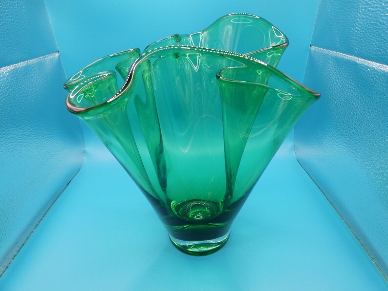 May include: A vibrant green glass vase with a ruffled rim and a flared, sculptural design. The vase has a rounded base and a wide opening, showcasing a modern aesthetic. The rim is accented with a thin gold trim.