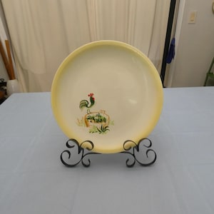 May include: A cream-colored ceramic plate with a yellow rim, featuring a rooster illustration. The rooster is green with a red comb, standing above a small planter with green foliage. The plate is displayed on a black metal stand.
