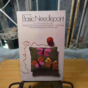 May include: A book titled "Basic Needlepoint" by Maxine Searls, displayed on a black metal stand. The cover features a small, embroidered pillow with colorful butterfly designs. A ball of yarn sits near the top of the book.