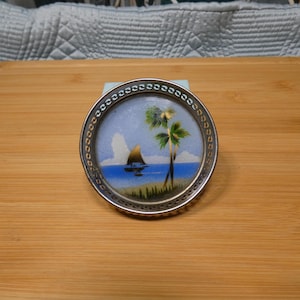 May include: A small, round coaster with a silver-colored metal rim. The coaster features a painted scene of a sailboat on blue water, a palm tree, and a light blue sky. The coaster is likely made of glass or a similar material.