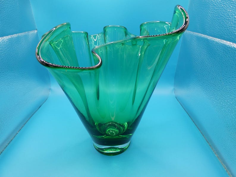 May include: A green glass vase with a ruffled rim and a silver-colored trim. The vase has a flared shape and a rounded base. The glass is transparent, allowing light to pass through. The vase is a decorative item.