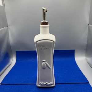 May include: White ceramic olive oil bottle with a stainless steel spout and cork stopper. The bottle has a curved shape and features the words "Olio d'oliva" and an olive branch design. The base of the bottle has the words "Olio Carli".