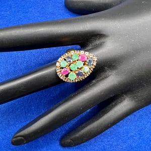 May include: A gold-toned ring with an oval cluster of colorful gemstones. The ring features a mix of green, pink, blue, and clear stones, set in a detailed, textured band. The ring is displayed on a black surface against a blue background.