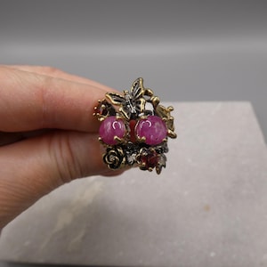 May include: A detailed ring featuring two large, round, pink gemstones. The ring has an ornate design with gold and silver accents, including a butterfly, roses, and smaller red gemstones. The ring is held in a hand.