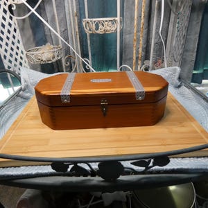 May include: A wooden box with a hinged lid and metal accents. The box is a warm brown color and has two silver straps across the top. The box is sitting on a wooden cutting board.