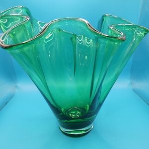 May include: A vibrant green glass vase with a ruffled rim and a silver-colored trim. The vase has a flared, organic shape and sits on a clear base. The vase is approximately 10 inches tall.