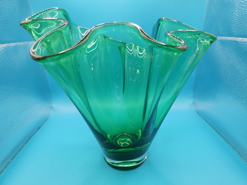 May include: A vibrant green glass vase with a ruffled rim and a silver-colored trim. The vase has a flared, organic shape and sits on a clear base. The vase is approximately 10 inches tall.