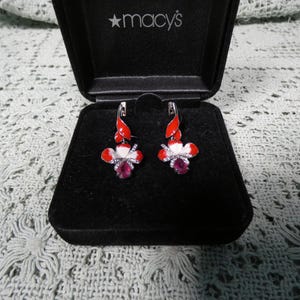 May include: A pair of flower-shaped earrings in a black jewelry box. The earrings feature red and white enamel petals, a central pink gemstone, and small clear crystals. The earrings are displayed in a black box with the Macy's logo.