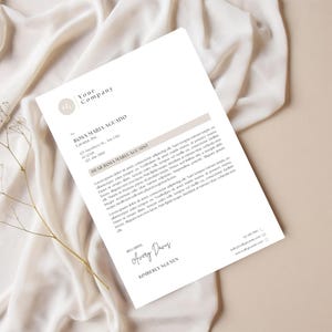 以下が含まれることがあります： 「Your Company」と「ROSA MARIA AGUADO」のテキストがミニマリストスタイルで印刷された白いレターヘッド。 レターヘッドは、ドライフラワーの小枝が付いたベージュの生地の上に置かれています。