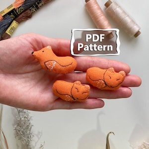 以下が含まれることがあります： 手の中に、刺繍が施されたオレンジ色のキツネの形をした3つの布製オーナメント。画像には、さまざまな色の糸巻きと小さな黄色のカボチャも写っています。「PDF Pattern」と書かれたサイン。