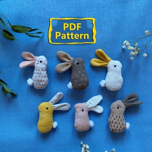 May include: Six handmade fabric bunny ornaments in various colors, including white, yellow, pink, and gray, with contrasting ear colors. The bunnies are arranged on a blue background with the text "PDF Pattern".