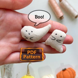 Könnte beinhalten: Zwei kleine, cremeweiße Stoffgespenster mit gestickten schwarzen Augen und Mündern. Über einem Gespenst befindet sich eine Sprechblase mit dem Text "Boo!". Ein rechteckiges orangefarbenes Etikett mit schwarzem Text zeigt "PDF Pattern".
