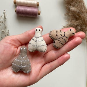 May include: Three small, handmade fabric dolls in beige and off-white, with black stitched details. The dolls are held in a hand, with two spools of thread in the background. The dolls have a simple, abstract design.