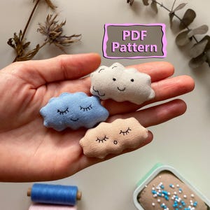 May include: Three small, cloud-shaped fabric ornaments in light blue, beige, and off-white, each with embroidered eyes and smiles. The image also shows spools of thread and a container with beads. The text "PDF Pattern" is visible.