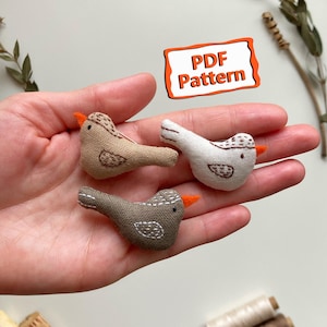 Op de afbeelding: Drie kleine, handgemaakte stoffen vogelornamenten in bruin- en wittinten, met oranje snavels en geborduurde details. De vogels worden in een hand gehouden, met de tekst "PDF Pattern" in een oranje kader. Naaibenodigdheden op de achtergrond.