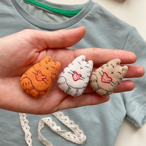 May include: Three small, handmade cat-shaped fabric ornaments in orange, white, and tan, each with a heart design. The cats are held in a hand, with a light blue top and lace trim in the background. Spools of thread are also visible.