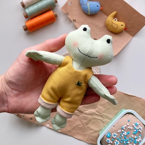 May include: A handmade frog doll with a yellow jumpsuit and green limbs is held in a hand. Two small felt bird ornaments and spools of thread in pastel colors are also visible. A pin cushion with blue and white pins is in the corner.