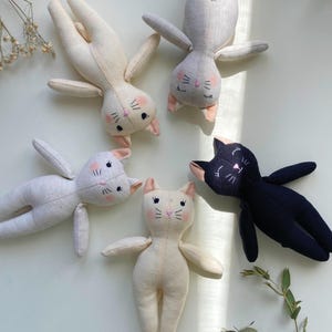 May include: Five handmade cat dolls in various colors. The dolls are made of soft fabric and feature embroidered facial details, including pink cheeks, whiskers, and closed eyes. One doll is black, while the others are cream and gray.