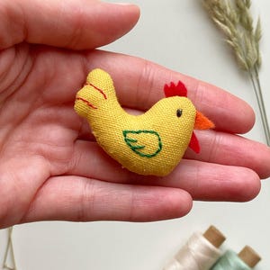 May include: A small, yellow fabric chicken with red and orange accents. The handmade chicken has a green embroidered design on its body. The chicken is held in a person's hand, with spools of thread in the background.