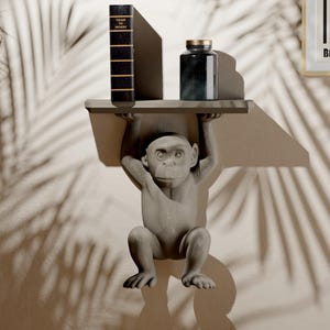 May include: A gray monkey-shaped shelf holds a book and a dark glass jar with a gold lid. The shelf is mounted on a wall, with shadows from a leafy plant. The shelf is a unique decorative item.