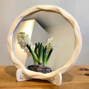 Könnte beinhalten: Ein runder, cremefarbener Spiegel mit gewelltem Rahmen, der von einem dekorativen Ständer getragen wird. Der Spiegel reflektiert eine weiße Wand und einen Blick in einen Raum. Vor dem Spiegel steht eine Topfhyazinthe mit weißen Blüten und grünen Blättern.