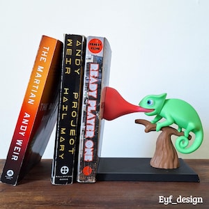 May include: A whimsical bookend featuring a green chameleon with a long red tongue, perched on a brown branch. The bookend is holding up three books: "The Martian", "Project Hail Mary", and "Ready Player One". The bookend is on a black base.