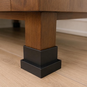 May include: A matte black square furniture leg riser featuring a stepped, tiered design. The riser is fitted securely around the base of a thick, square-shaped wooden furniture leg, providing a stable and elevated base for the furniture piece.