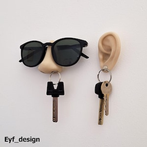 Può includere: Un set di due ganci per chiavi, uno a forma di naso con occhiali da sole e l'altro a forma di orecchio. Entrambi i ganci hanno chiavi appese.