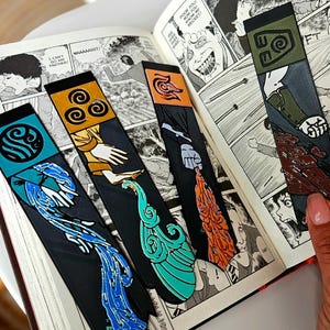 May include: Four black bookmarks with colorful designs are inserted into an open manga book. Each bookmark features a different design: a blue water symbol, a yellow swirl, an orange face, and a green swirl. The book is open to a black and white comic.