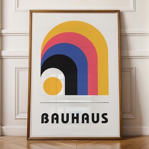 Könnte beinhalten: Ein gerahmter Bauhaus-Kunstdruck mit einem farbenfrohen geometrischen Design. Der Druck zeigt einen gelben, rosa, blauen und schwarzen Bogen, mit dem Wort "BAUHAUS" in schwarzen Blockbuchstaben. Der Rahmen ist hellbraun.