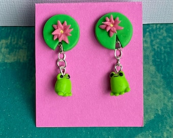 May include: A pair of handmade earrings featuring a green lily pad with a pink flower and a dangling green frog charm. The earrings are attached to a pink card.